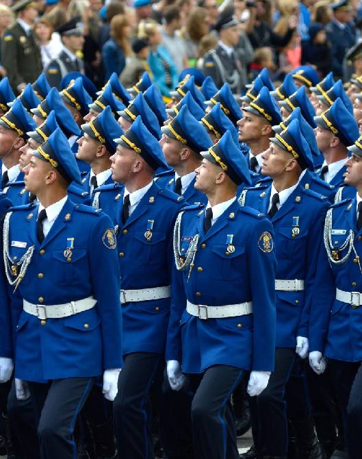 An honor guard marching in formation in dark ceremonial uniforms with gold braid, sashes, and peaked caps